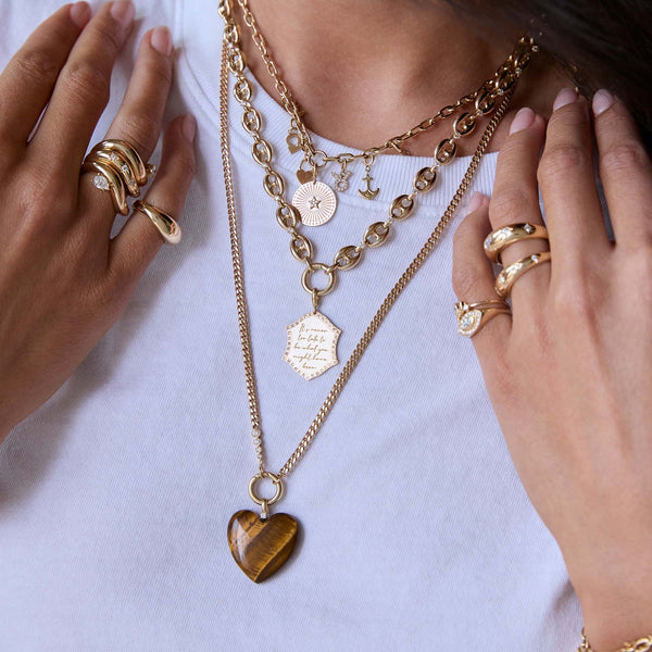 Close-up of a neck and fingers wearing Zoe Chicco 14k gold charm necklaces, featuring a tiger eye heart-shaped pendant and diamond rings against a white t-shirt
