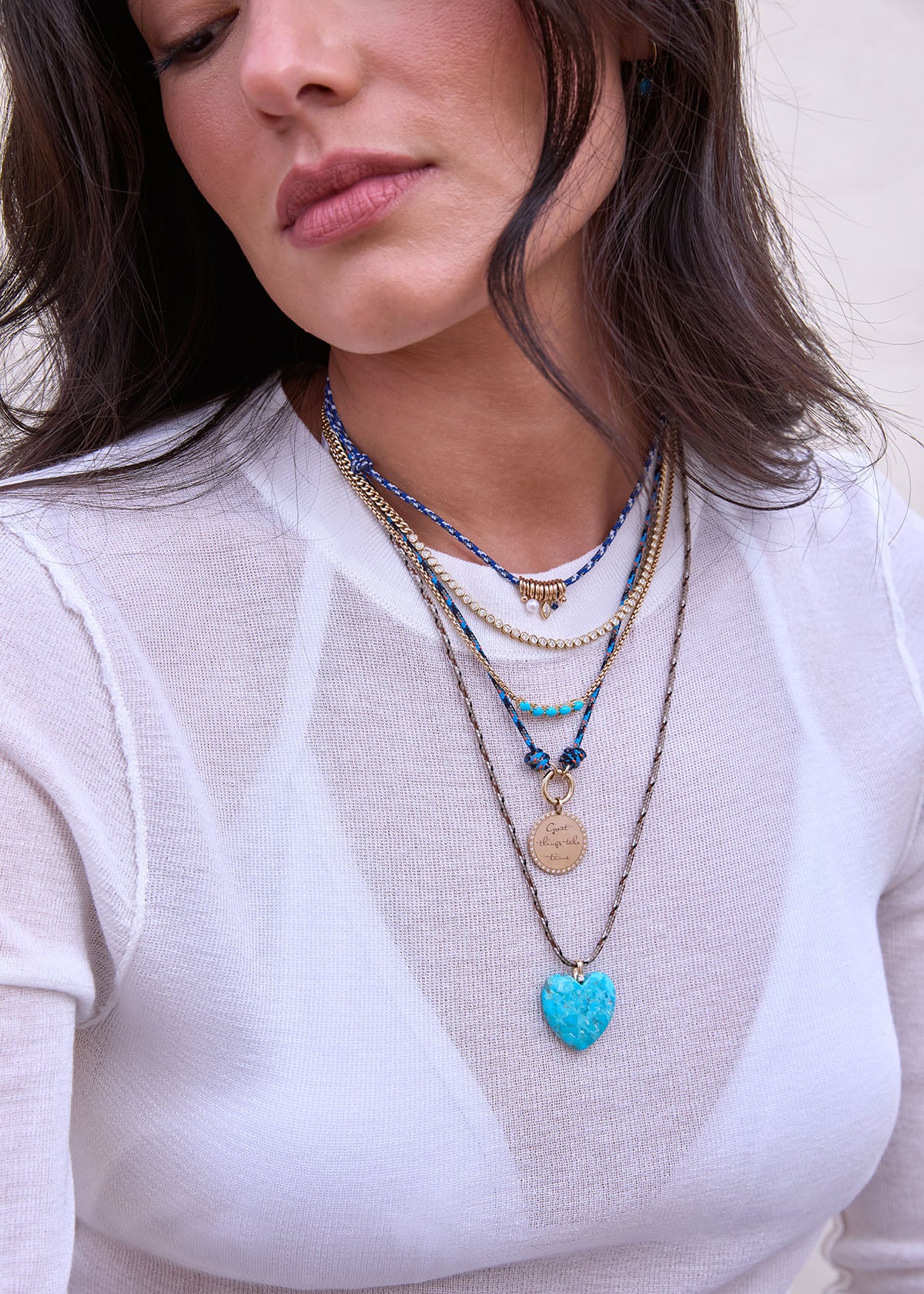 Woman wearing a white shirt with multiple Zoe Chicco 14k gold paracord necklaces with a blue heart pendant and turquoise and diamond tennis