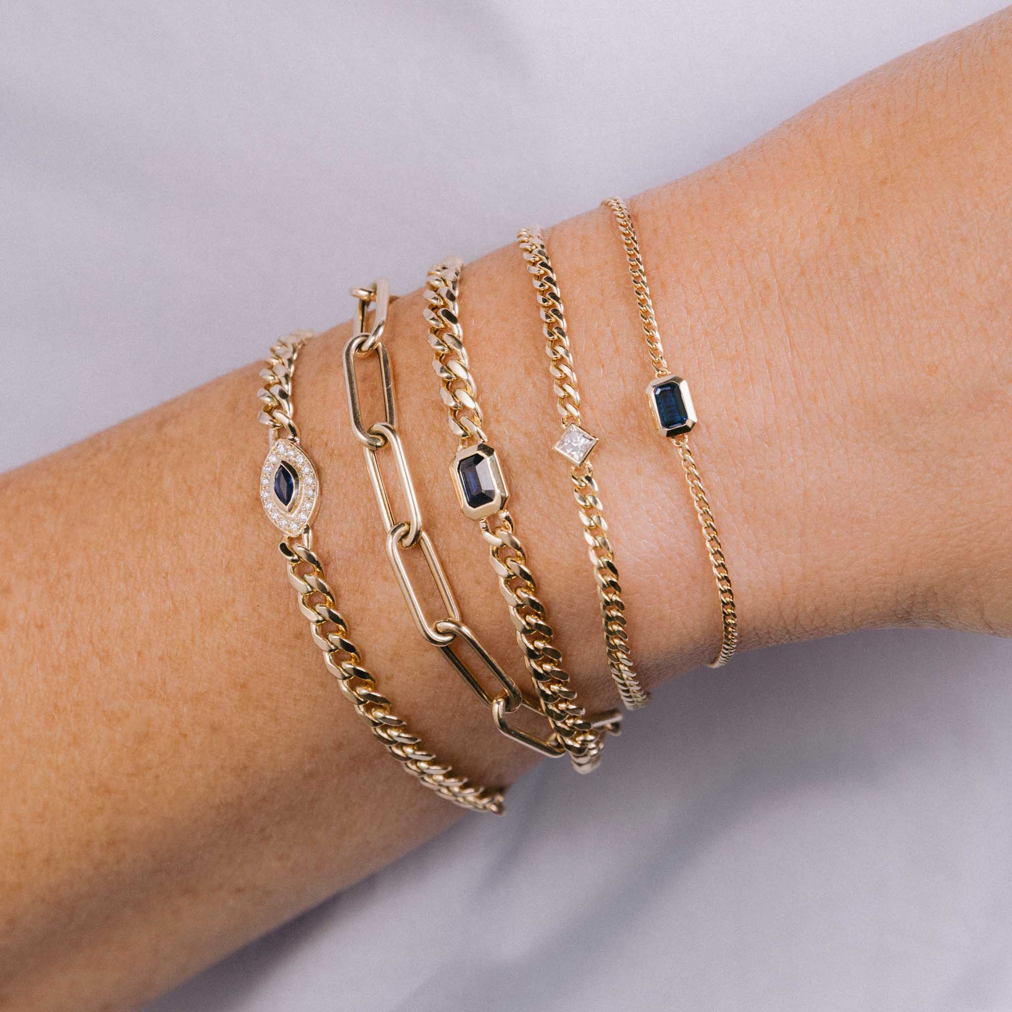 close up of a wrist wearing a Zoë Chicco 14k Gold Medium Curb Chain Emerald Cut Blue Sapphire Bezel Bracelet stacked with a 14k Medium Curb Chain Marquise Blue Sapphire Halo Bracelet, Large Paperclip Chain Bracelet, 14k Princess Diamond Small Curb Chain Bracelet, and a 14k Emerald Cut Blue Sapphire XS Curb Chain Bracelet