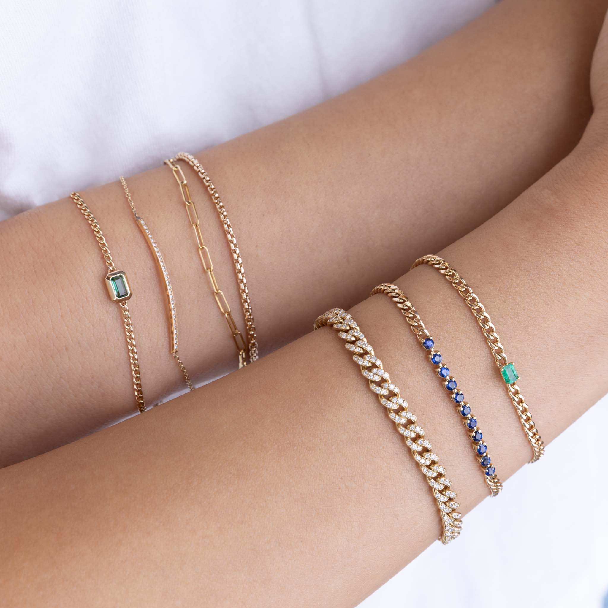 a woman's wrist wearing a Zoë Chicco 14k Gold Small Box Chain Bracelet stacked with a Small Paperclip Chain Bracelet, 14k Emerald Cut Emerald XS Curb Chain Bracelet, and a 14k Pavé Diamond Thin Bar Bracelet next to another wrist wearing a 14k Emerald Cut Emerald Small Curb Chain Bracelet stacked with a Blue Sapphire Tennis Segment Curb Chain bracelet and a Pavé Diamond Medium Curb Chain Bracelet