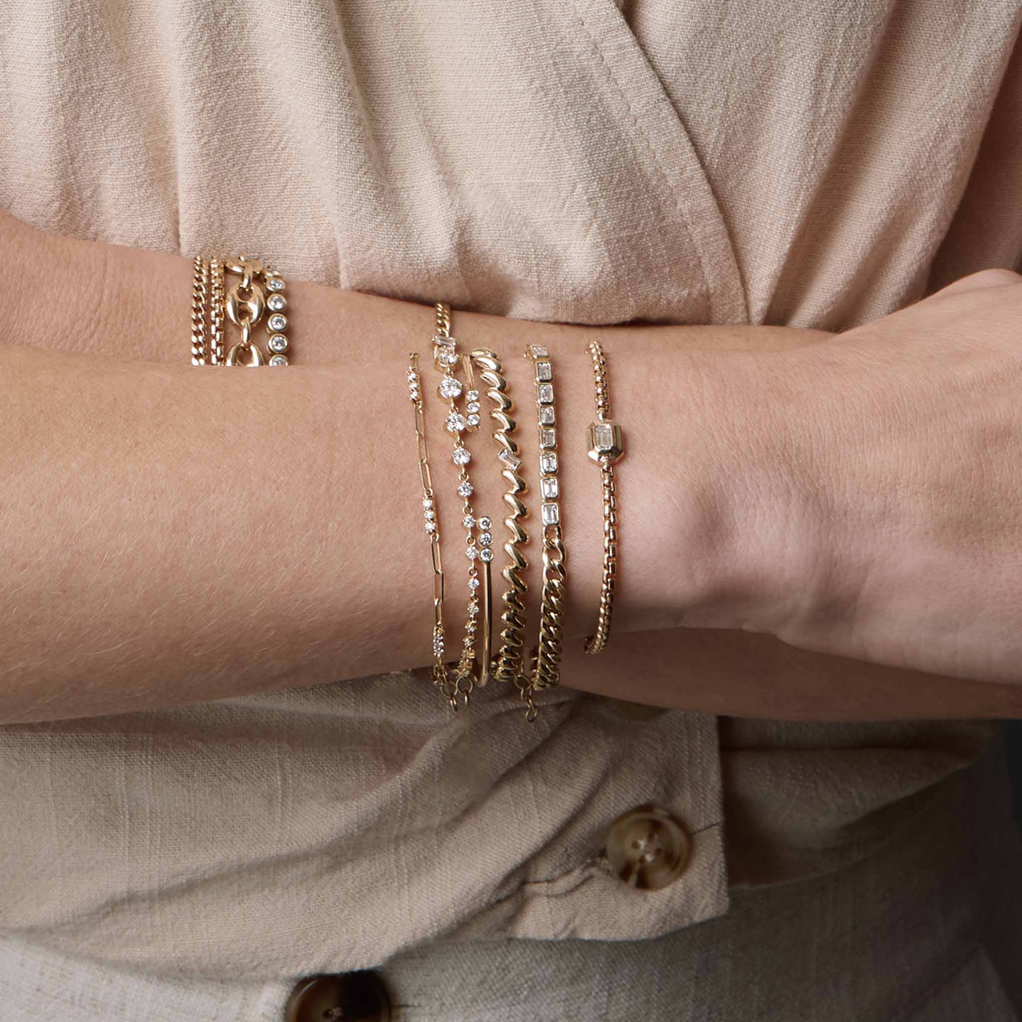 woman's wrist wearing a Zoë Chicco 14k Gold Emerald Cut Diamond Medium Box Chain Bracelet layered with a 14k Curb Chain & Emerald Cut Diamond Half Tennis Bracelet, Baguette Diamond San Marco Chain Bracelet, 14k 3 Diamond Bezel Bar Open Cuff, 14k Linked Graduated Prong Diamond Tennis Bracelet, and a 14k Prong Diamond Bar Station Small Paperclip Chain Bracelet