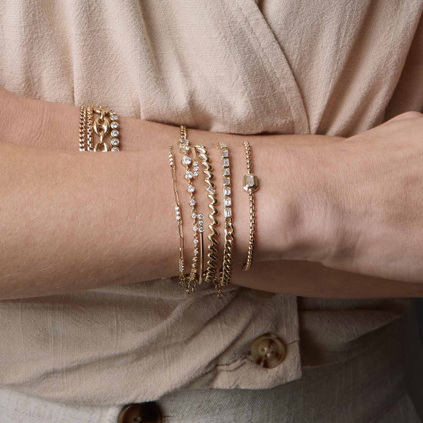 woman's wrist wearing a Zoë Chicco 14k Gold Emerald Cut Diamond Medium Box Chain Bracelet layered with a 14k Curb Chain & Emerald Cut Diamond Half Tennis Bracelet, Baguette Diamond San Marco Chain Bracelet, 14k 3 Diamond Bezel Bar Open Cuff, 14k Linked Graduated Prong Diamond Tennis Bracelet, and a 14k Prong Diamond Bar Station Small Paperclip Chain Bracelet