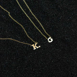 On black sand lies two necklaces:  a letter necklace with bezel set diamond shown as "K"  and a plain 14k gold block letter shown as "G".