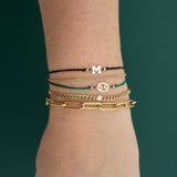  A woman wears the 14k Midi Bitty Zodiac Cord Bracelet in green and the 14k Letter Cord Bracelet in black with a small curb chain bracelet with floating diamond and a a large paperclip chain bracelet.