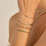 Close-up of a wrist wearing multiple Zoee Chicco 14k gold bracelets with diamonds and emeralds on a beige background