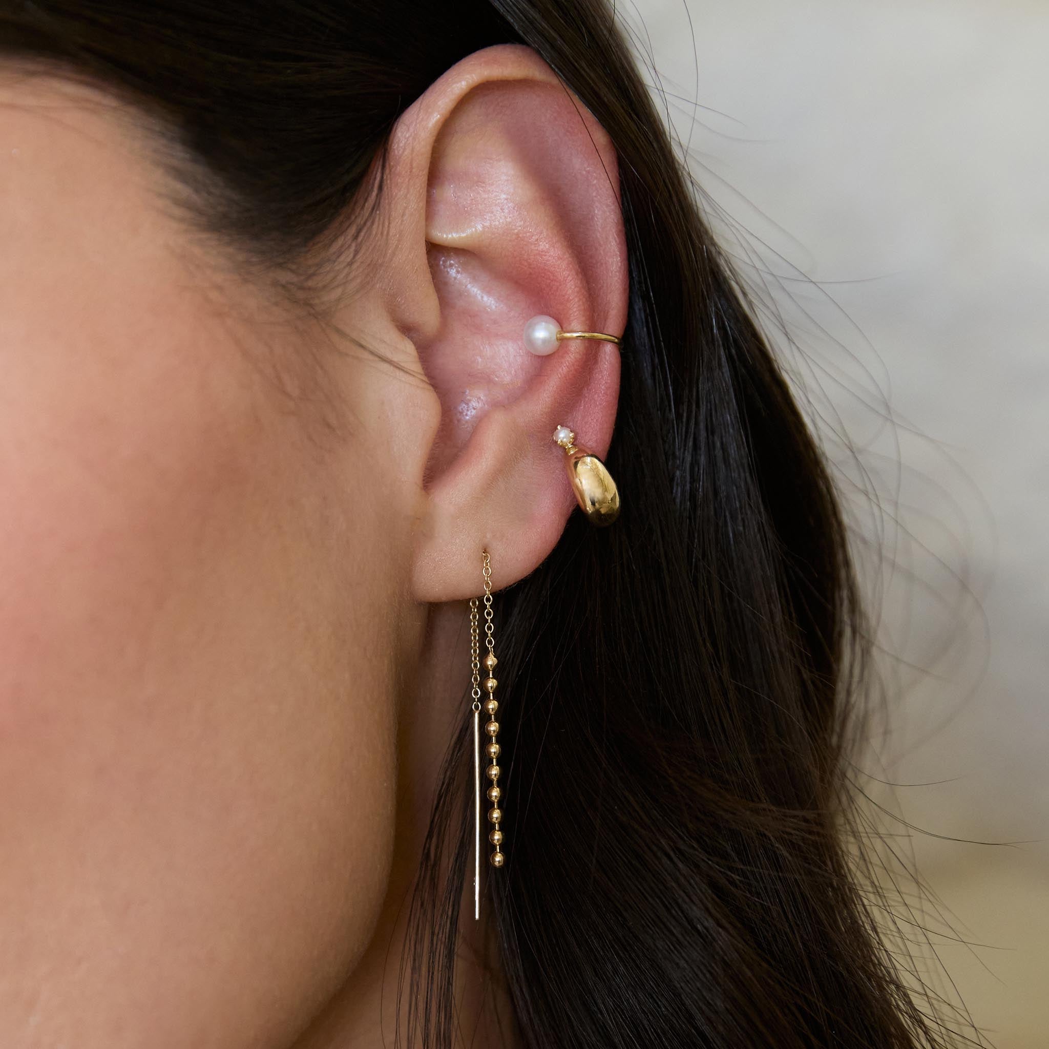 Close-up of an ear wearing Zoe Chicco 14k gold hoop earring with small pearl, bead threader earring, and a white pearl ear cuff on a neutral background