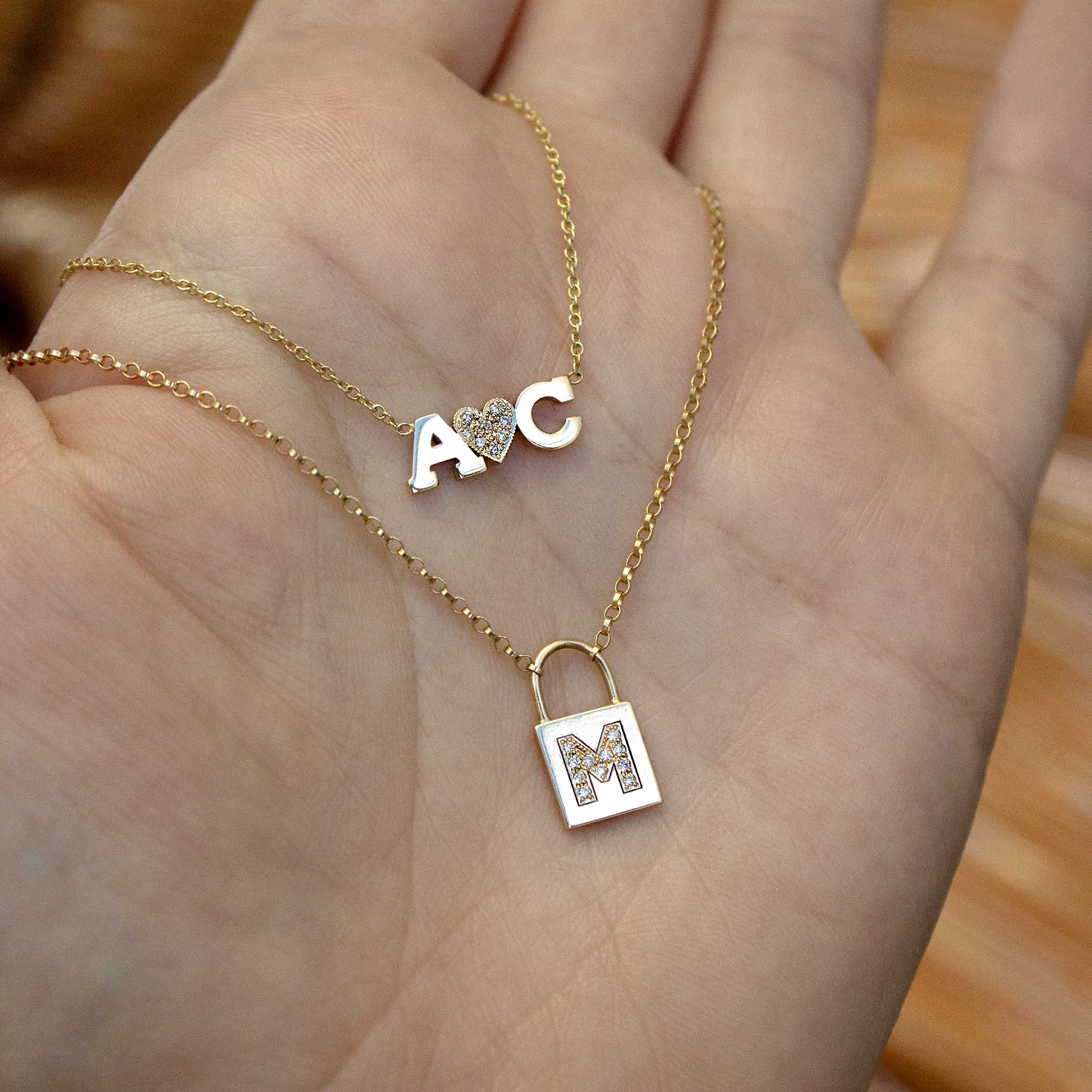 In a woman's hand are two 14k gold necklaces including a 2-block letter with pave diamond heart necklace and a pave letter padlock necklace.