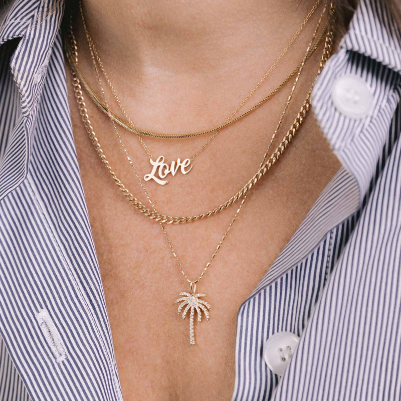 A woman wears Zoe Chicco's 14k Gold Script Letter Love Necklace, Snake and small curb chains and a pave diamond covered palm tree necklace