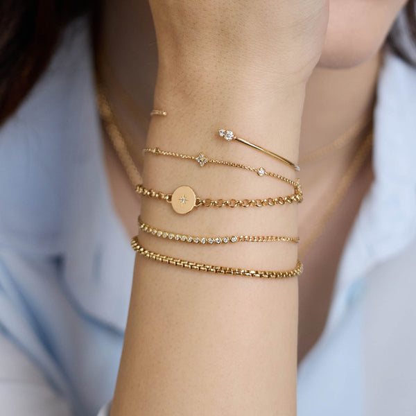 Close-up of a woman's wrist wearing multiple Zoe Chicco 14k gold and diamond chain bracelets with various designs with a blurred light blue background