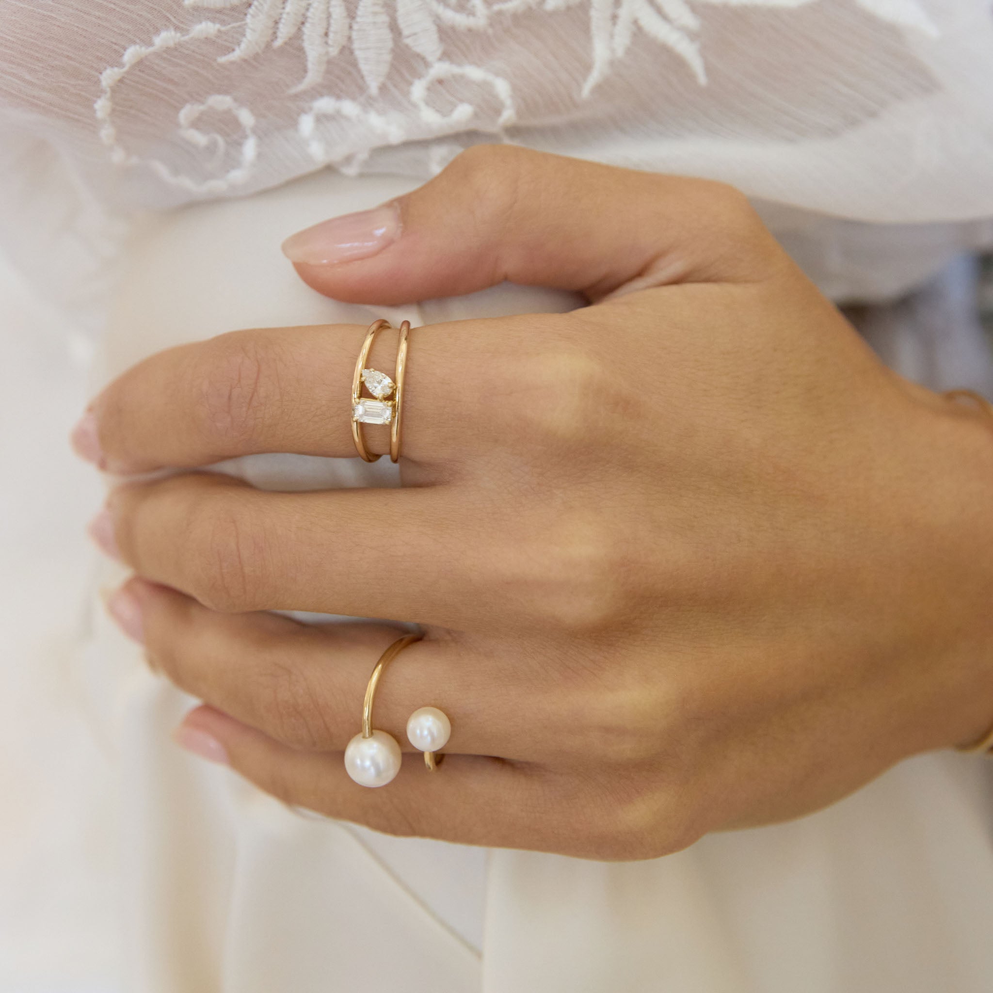 Hand wearing Zoe Chicco 14k gold ring with emerald cut and pear diamonds and a pearl wrap ring against a white lace background
