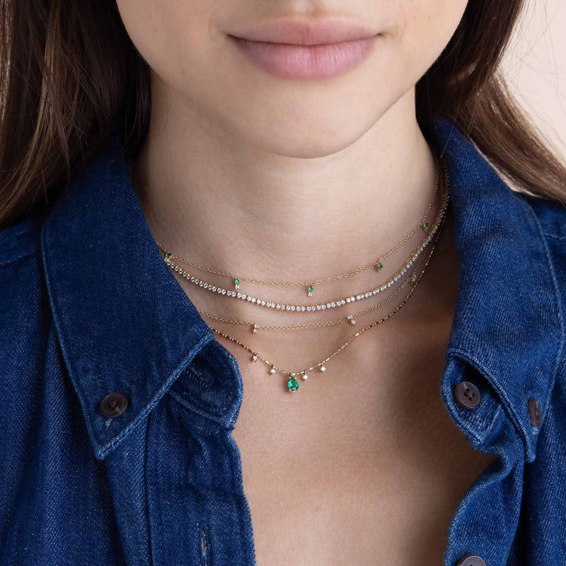 woman in a denim shirt wearing a Zoë Chicco 14k Gold Pear Emerald Pendant & Dangling Diamonds Tube Bar Chain Necklace layered with a 14k Stacked Prong Emerald & Diamond Station Necklace and a Stacked Prong Diamond Station Necklace