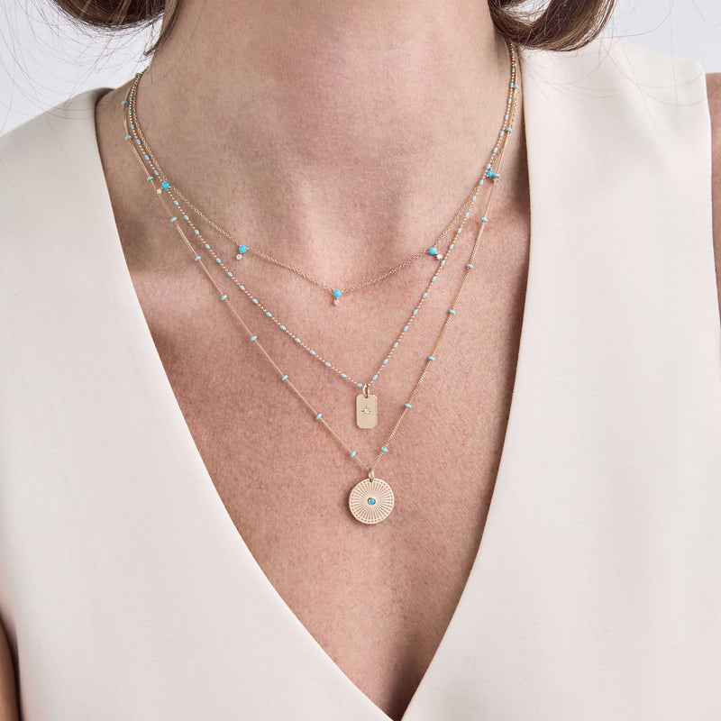 woman in a beige vest wearing a Zoë Chicco 14k Gold Star Set Diamond Small Square Edge Dog Tag Necklace on a Turquoise Enamel Tube Bar Chain layered with a Stacked Turquoise & Prong Diamond Station necklace and a 14k Gold Turquoise Sunbeam Medallion Satellite Chain Necklace