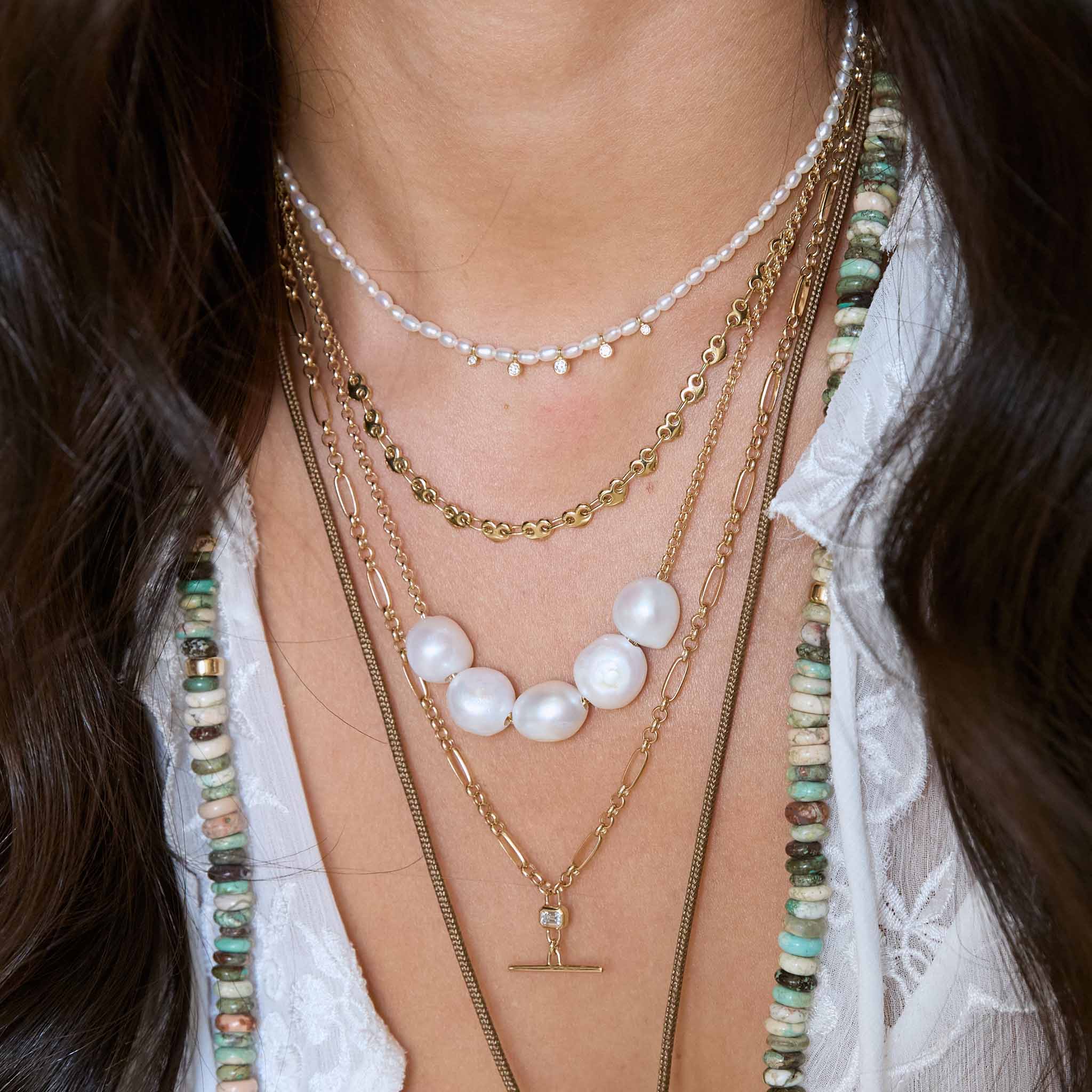 Close-up of a women's neck wearing Zoe Chicco 14k gold chain necklaces with pearls and diamonds and green gemstones wearing a white lace blouse