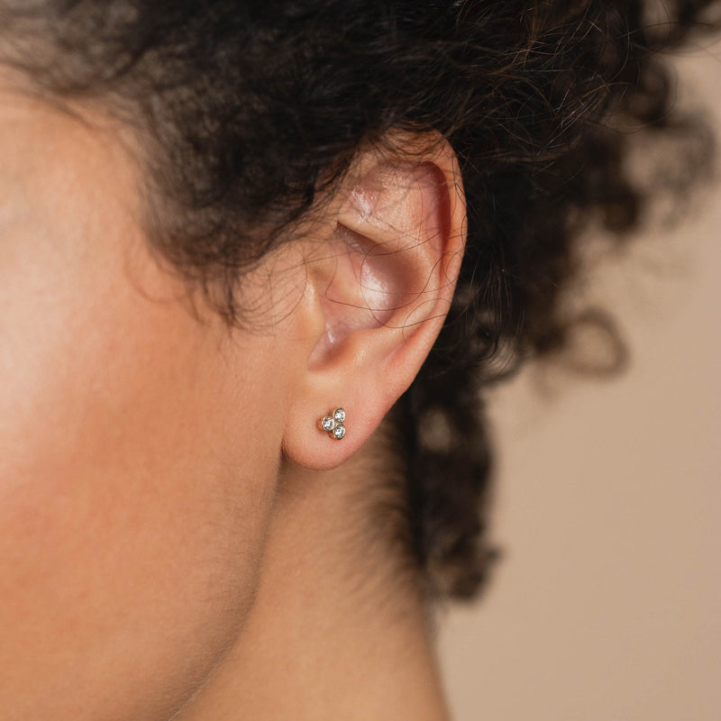 A woman with dark hair is wearing a trio of bezel diamond stud earrings