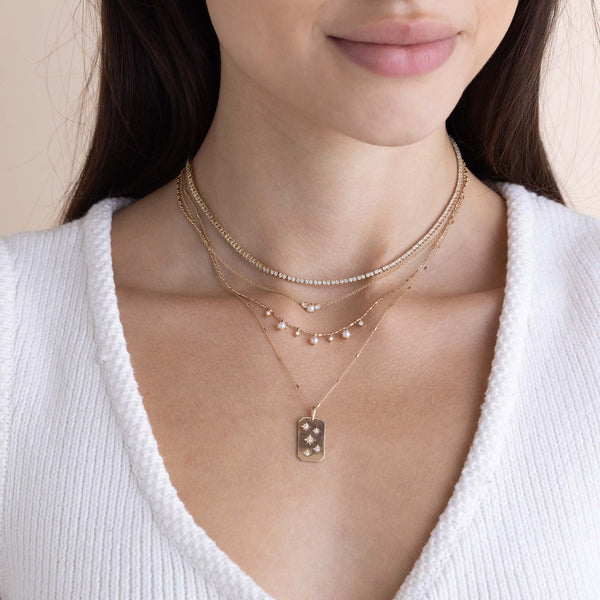 a woman in a white sweater wearing a Zoë Chicco 14k Gold 7 Dangling Prong Pearl & Diamond Tube Bar Chain Necklace layered with a 14k Prong Diamond Tennis Necklace and a 14k Scattered Star Set Diamonds Square Edge Dog Tag Necklace