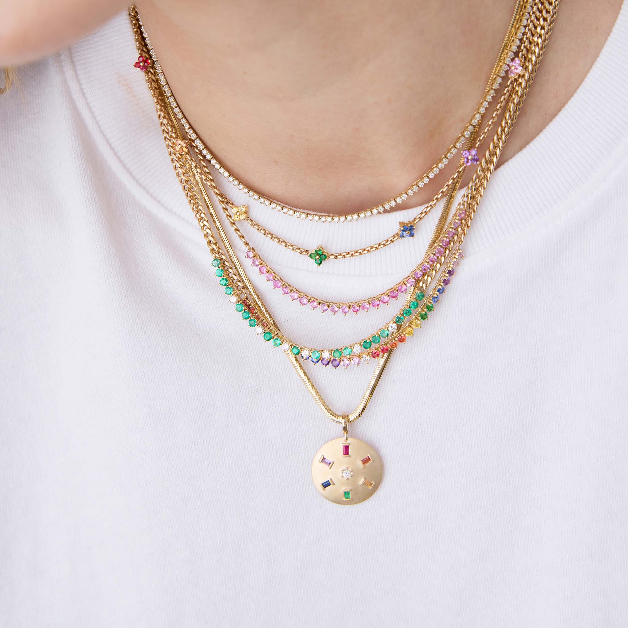 woman in a white tshirt wearing a Zoë Chicco 14k Gold Rainbow Sapphire Tennis Segment Small Curb Chain Necklace layered with a 14k Pink Sapphire Tennis Segment Small Curb Chain Necklace, 14k Emerald & Diamond Tennis Segment Small Curb Chain Necklace, 14k Ruby Tennis Segment Small Curb Chain Necklace, 14k Rainbow Sapphire Quad Station Box Chain Necklace, Diamond Tennis Necklace, and a 14k Single Rainbow Baguette & Diamond Brushed Gold Domed Disc Pendant hanging of a snake chain