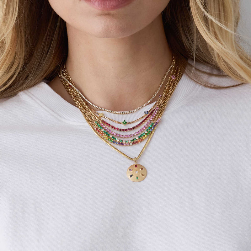 woman in a white tshirt wearing a Zoë Chicco 14k Gold Rainbow Sapphire Tennis Segment Small Curb Chain Necklace layered with a 14k Pink Sapphire Tennis Segment Small Curb Chain Necklace, 14k Emerald & Diamond Tennis Segment Small Curb Chain Necklace, 14k Ruby Tennis Segment Small Curb Chain Necklace, 14k Rainbow Sapphire Quad Station Box Chain Necklace, Diamond Tennis Necklace, and a 14k Single Rainbow Baguette & Diamond Brushed Gold Domed Disc Pendant hanging of a snake chain