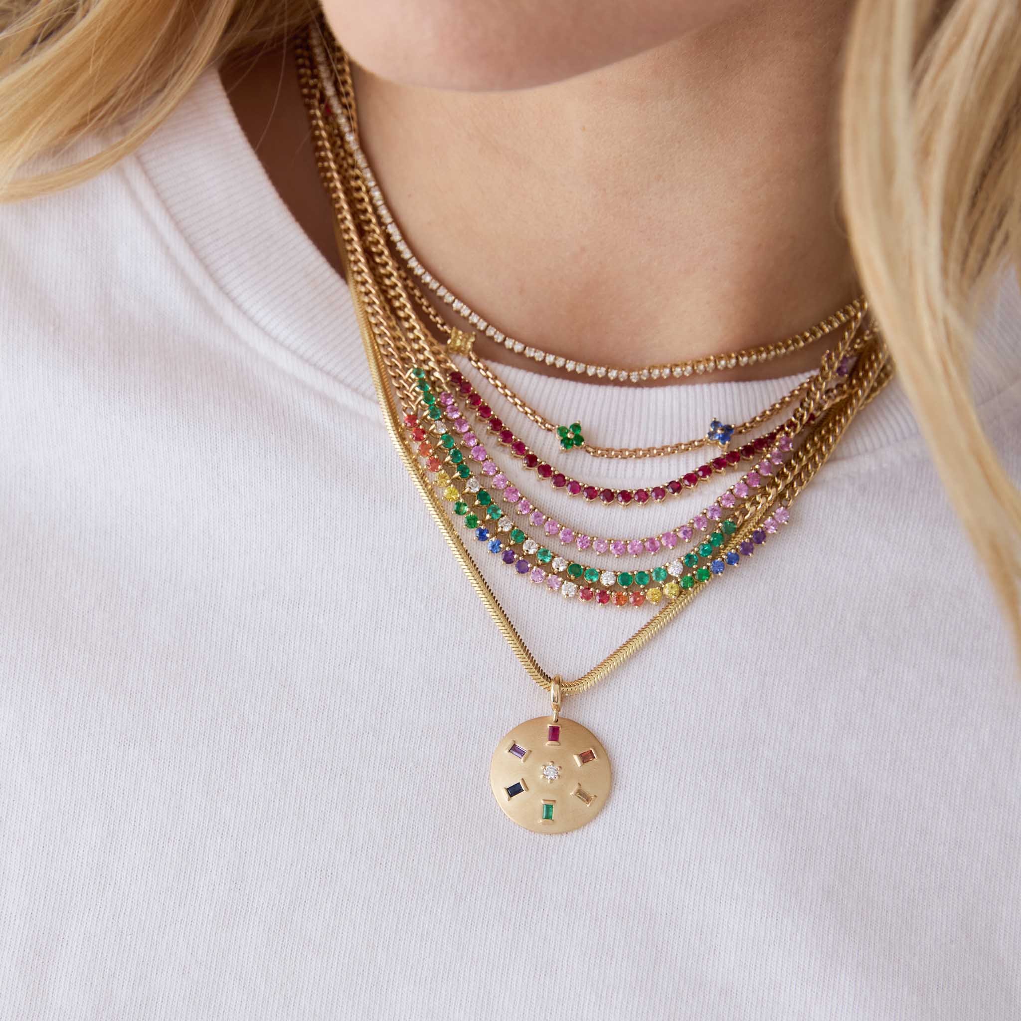 woman in a white tshirt wearing a Zoë Chicco 14k Gold Rainbow Sapphire Tennis Segment Small Curb Chain Necklace layered with a 14k Pink Sapphire Tennis Segment Small Curb Chain Necklace, 14k Emerald & Diamond Tennis Segment Small Curb Chain Necklace, 14k Ruby Tennis Segment Small Curb Chain Necklace, 14k Rainbow Sapphire Quad Station Box Chain Necklace, Diamond Tennis Necklace, and a 14k Single Rainbow Baguette & Diamond Brushed Gold Domed Disc Pendant hanging of a snake chain