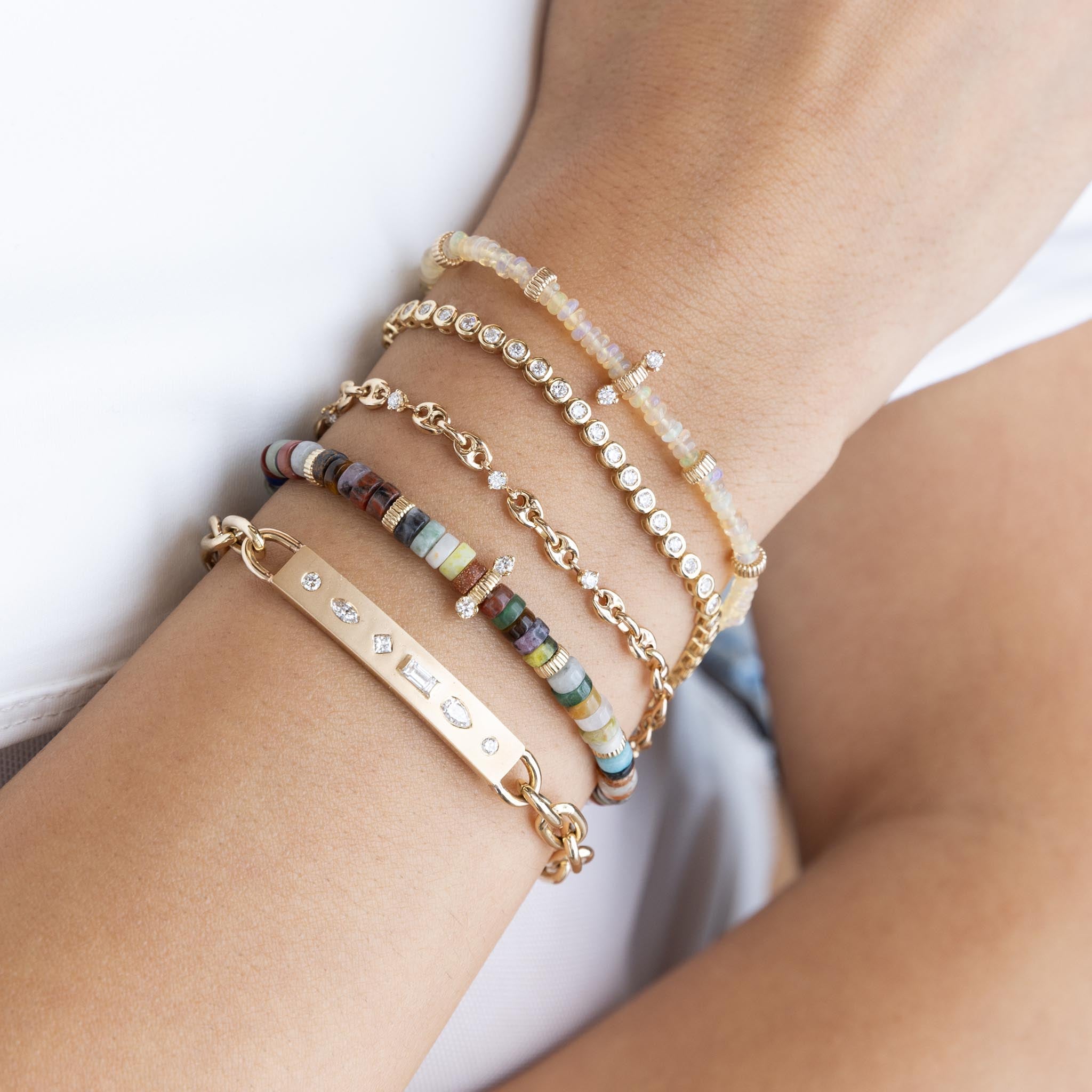 woman's wrist wearing a Zoë Chicco 14k Gold & Mixed Gemstone Heishi Bead Bracelet with 2 Prong Diamonds stacked with a 14k Diamond Mosaic Brushed Gold Horizontal ID Bar Bracelet, Fire Opal Rondelle Bead Bracelet with 2 Diamonds, Diamond Bezel Tennis Bracelet, and a Prong Diamond Small Mariner Chain Bracelet