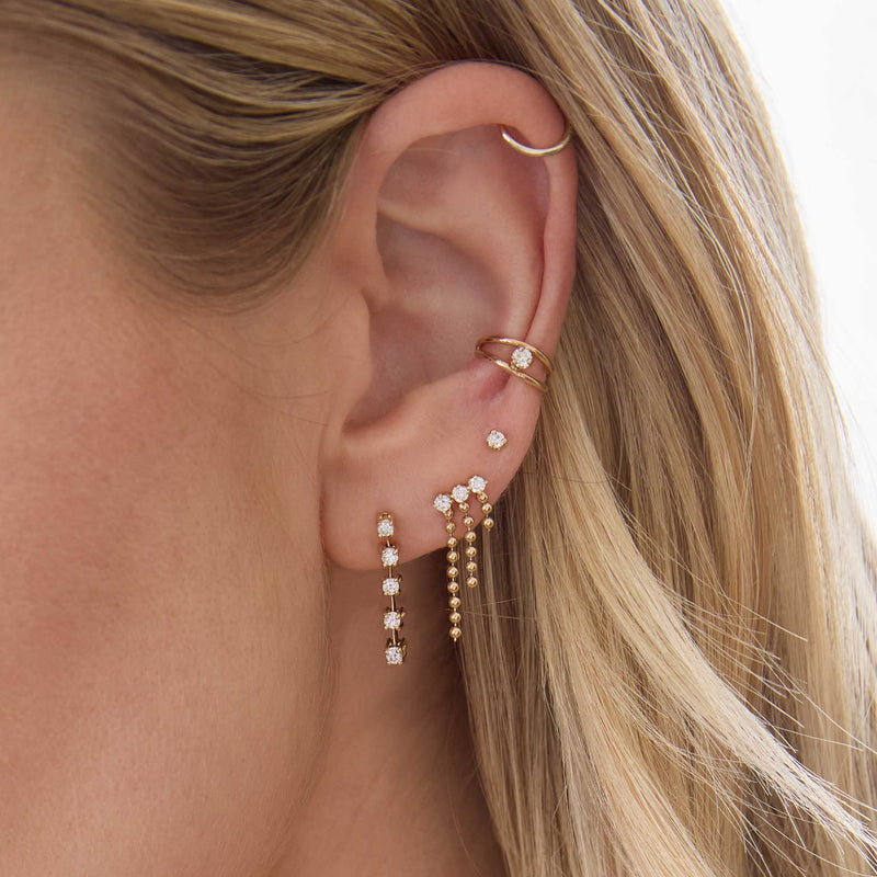 woman's ear wearing a Zoë Chicco 14k Gold Prong Diamond Dash Tennis Drop Earring layered with a 14k Prong Diamond  & Bead Chain Waterfall Earring, Prong Diamond Stud, and wearing a 14k Nested Prong Diamond Double Wire Ear Cuff