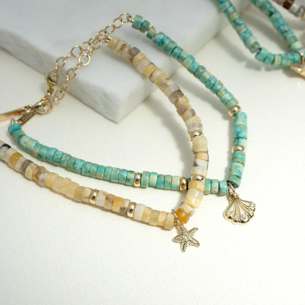 14k Gold & Sandy Feldspar Mixed Heishi Bead Starfish Charm Bracelet and 14k Gold & Light Turquoise Heishi Bead Seashell Charm Bracelet laying flat against a white background