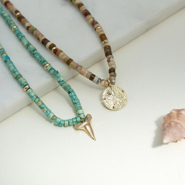 Zoe Chicco's 14k Gold & Light Turquoise Heishi Bead Shark's Tooth Charm Necklace and1 4k Gold & Sandy Feldspar Mixed Heishi Bead Sand Dollar Charm Necklace necklace shown on white.