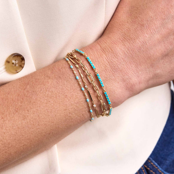 Close up of a woman's wrist wearing a Zoë Chicco 14k Gold & Turquoise Enamel Tube Bar Chain Bracelet layered with a Turquoise Bead Bar Bracelet, 14k Gold Bar & Graduated Prong Diamond Bracelet, and a 14k Medium Square Oval Chain Bracelet with 5 Pavé Diamond Links