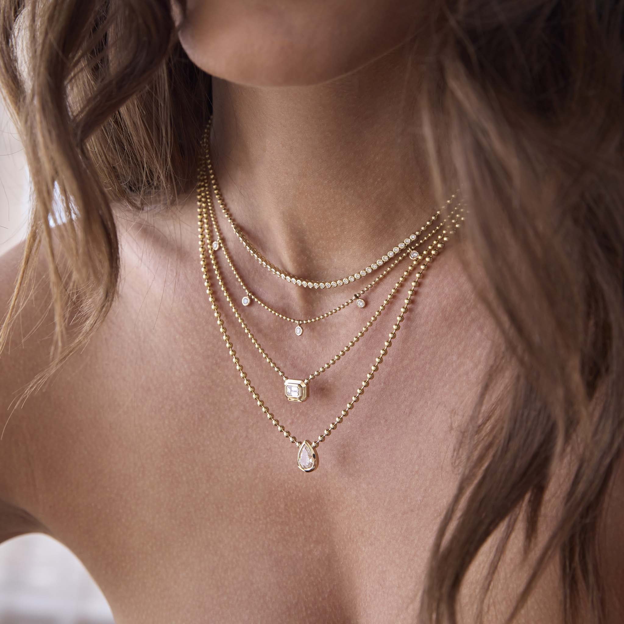 woman wearing a Zoë Chicco 14k Gold Large Emerald Cut Diamond Bezel Bead Chain Necklace layered with a 14k 5 Dangling Floating Diamond Station Bead Chain Necklace, one of a kind 1.03 ctw Mine Cut Pear Shaped Diamond Bead Chain Necklace, and a Diamond Bezel Tennis Segment Bead Chain Necklace