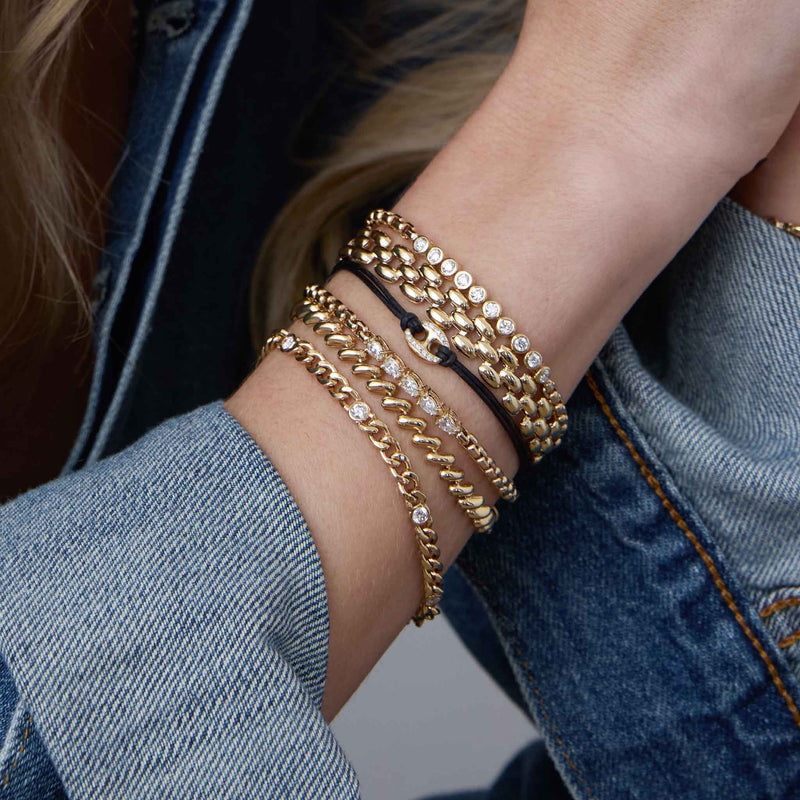 woman in a denim jacket wearing a Zoë Chicco 14k Gold Diamond Bezel Tennis Segment Large Box Chain Bracelet on her wrist stacked with a 14k Gold Medium Panther Chain Bracelet, 14k Large Pavé Diamond Mariner Link Leather Bracelet, 14k 5 Linked Pear Diamond Box Chain Bracelet, 14k Gold San Marco Chain Bracelet, and a 14k Medium Curb Chain Bracelet with 5 Floating Diamonds