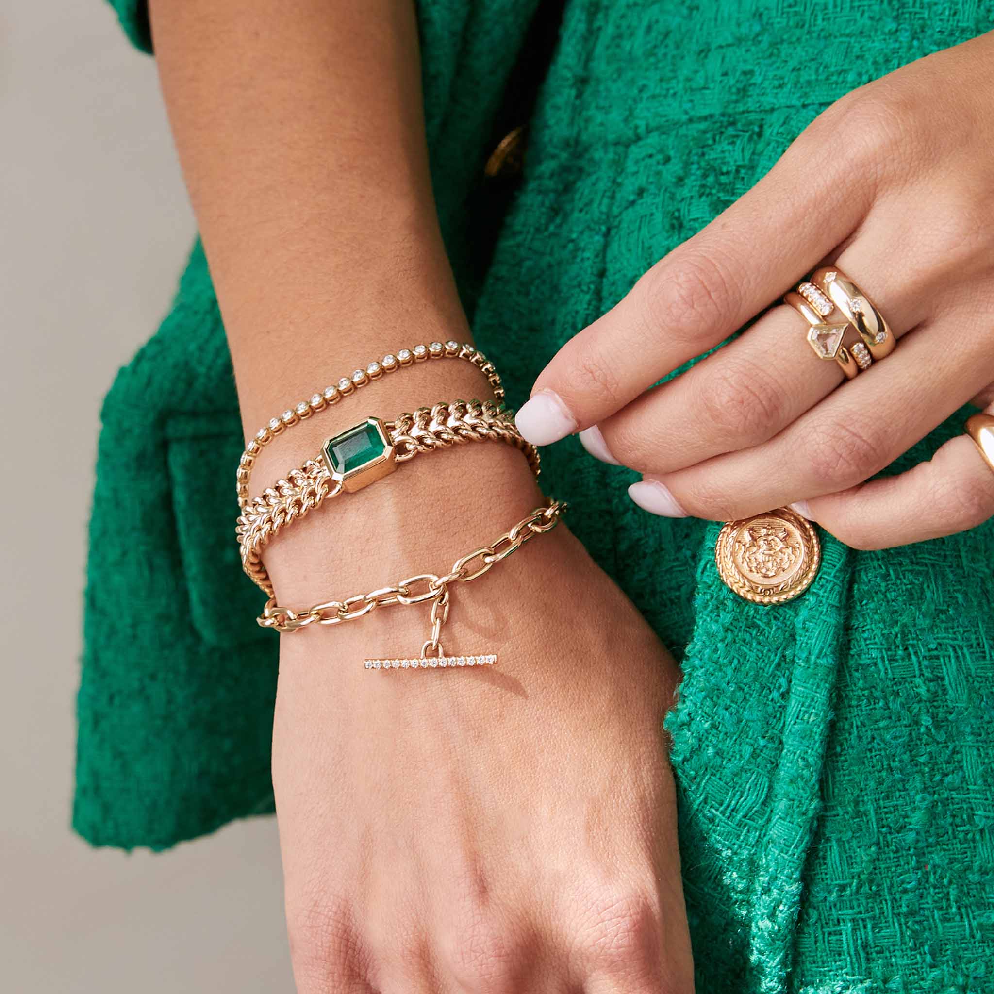 a close up of  a woman wearing a green tweed set wearing a Zoë Chicco 14k Gold One of a Kind Emerald Cut Emerald Bezel Double Wide Curb Chain Bracelet layered with two other bracelets on her wrist