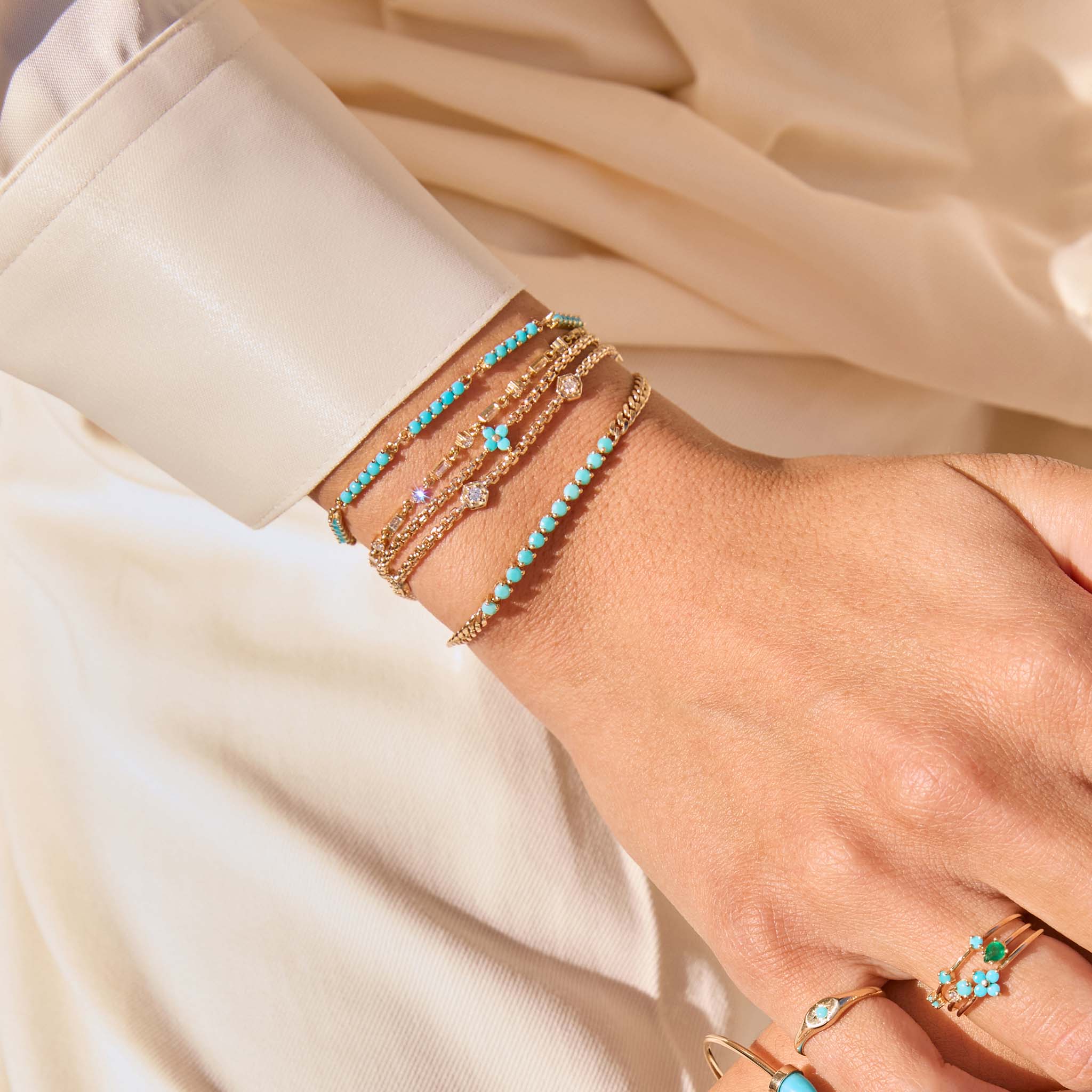 Close up of a wrist wearing multiple 14k gold chain bracelets with prong set diamonds and turquoise stones against white fabric