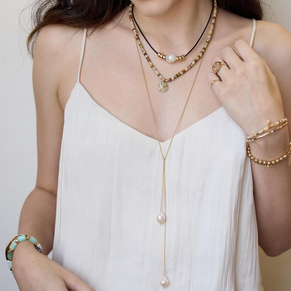 woman in a white camisole wearing a Zoë Chicco 14k Gold Baroque Pearl & Rondelle Bead Leather Cord Necklace layered with a 14k Gold & Sandy Feldspar Mixed Heishi Bead Sand Dollar Charm Necklace, and a 14k Double Baroque Pearl Wrap Around Lariat Necklace