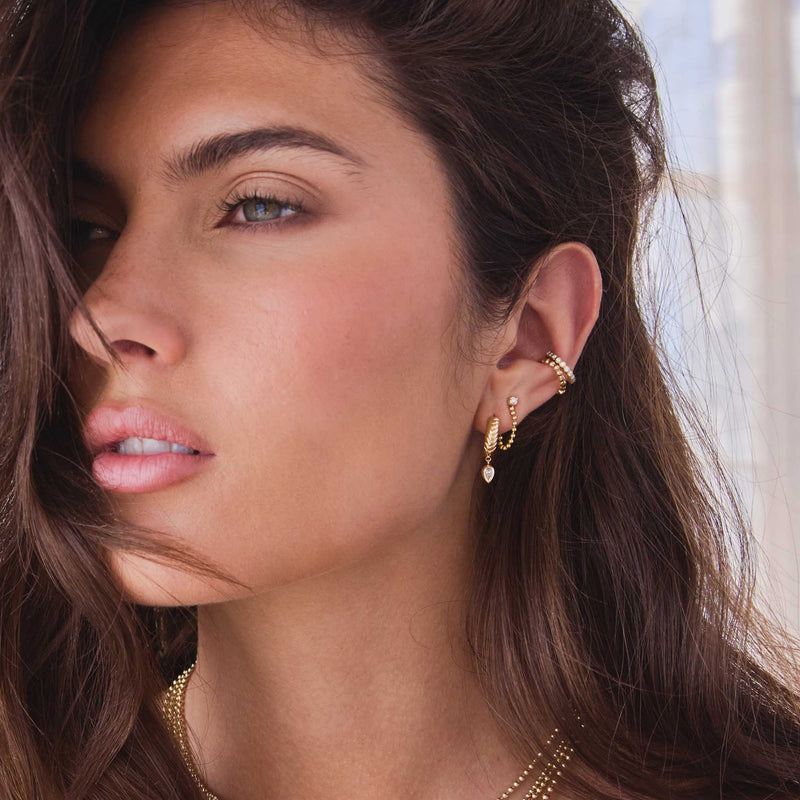 brunette woman wearing a Zoë Chicco 14k Gold Prong Diamond Bead Chain Huggie Earring in her second piercing layered with a 14k Pear Diamond Braided Hinge Huggie Hoop and two ear cuffs