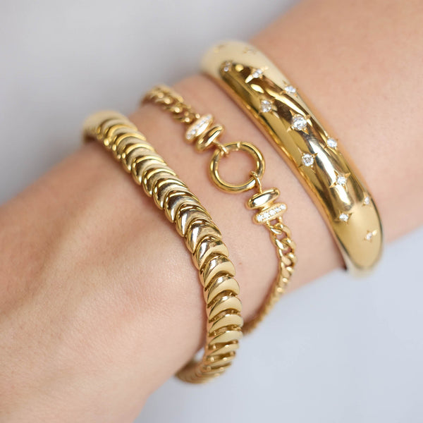 Close up of wrist wearing multiple Zoe Chicco 14k gold cuffs and bracelets on a blurred background