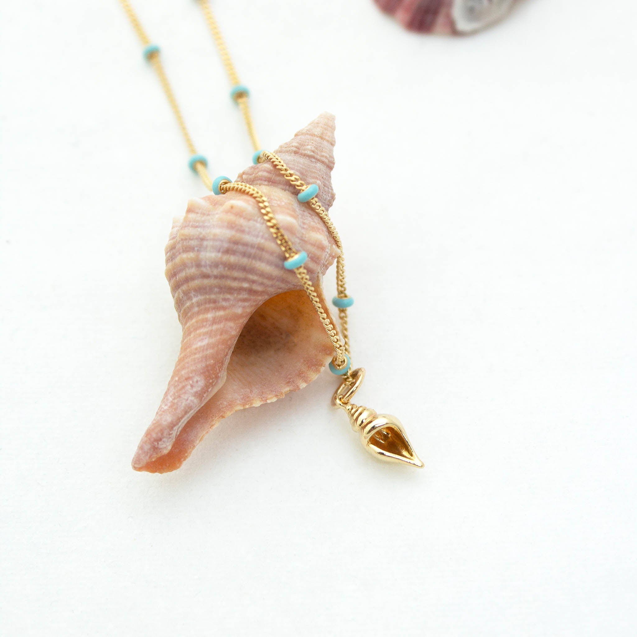 Zoë Chicco 14k Gold Conch Shell Turquoise Enamel Satellite Chain Necklace draped over a pink conch shell laying on a white background