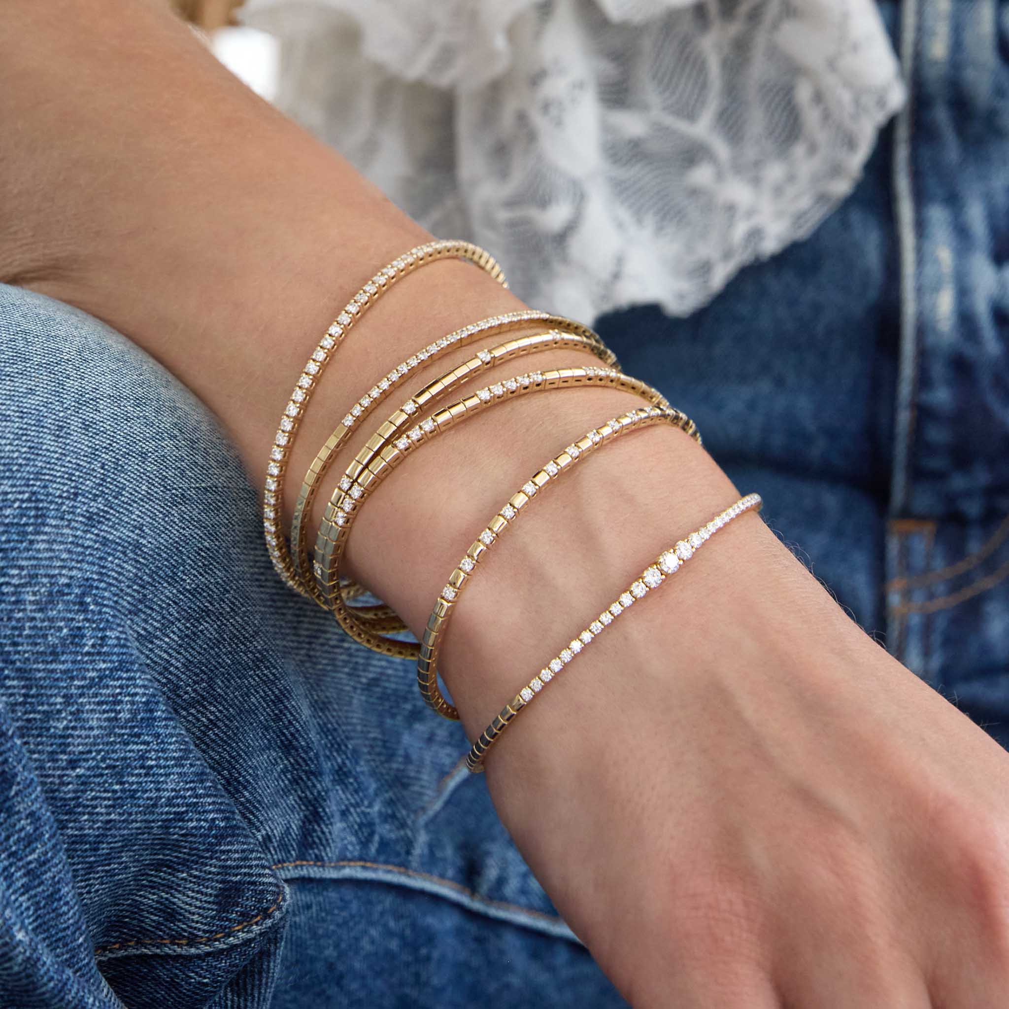 Close-up of a wrist wearing multiple Zoe Chicco 14k gold diamond bangle bracelets with a denim background