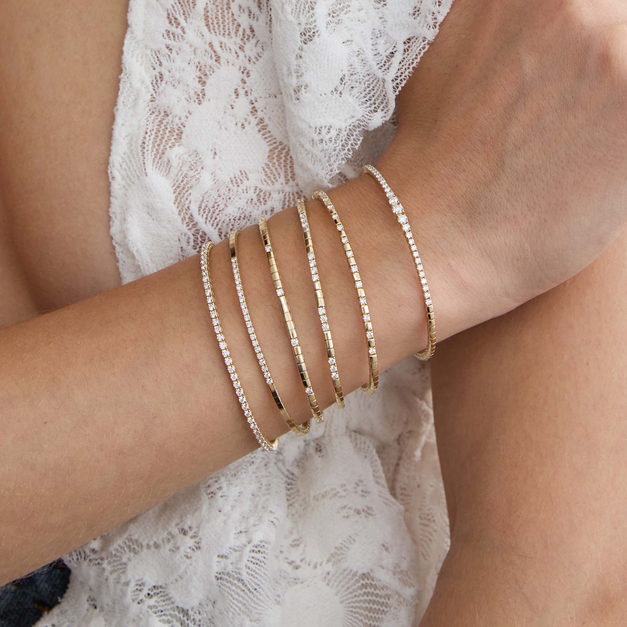 Close-up of a person's wrist wearing multiple Zoe Chicco 14k gold flex diamond tennis bangle bracelets on a light background