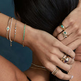 a woman holding her hands behind her head wearing a Zoë Chicco 14k Gold Emerald Tennis Segment Small Curb Chain Bracelet layered with three bracelets on her left wrist and various gold and gemstone rings stacked on her fingers