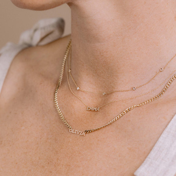close up of a woman wearing a Zoë Chicco 14k Gold Custom Itty Bitty Letters Curb Chain Necklace with the name OLIVIA layered with a Zoë Chicco 14k Gold Custom Itty Bitty Letters Necklace with the name EMMA and a Floating Diamond Station Necklace