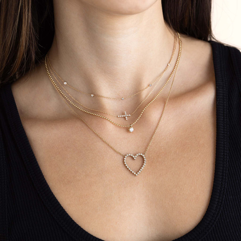 woman in a black top wearing a Zoë Chicco 14k Gold Small Diamond Bezel Cross Necklace layered with a 5 Floating Diamond Necklace, a 14k Prong Diamond Solitaire Bead Chain Necklace, and a 14k Diamond Bezel Heart Necklace