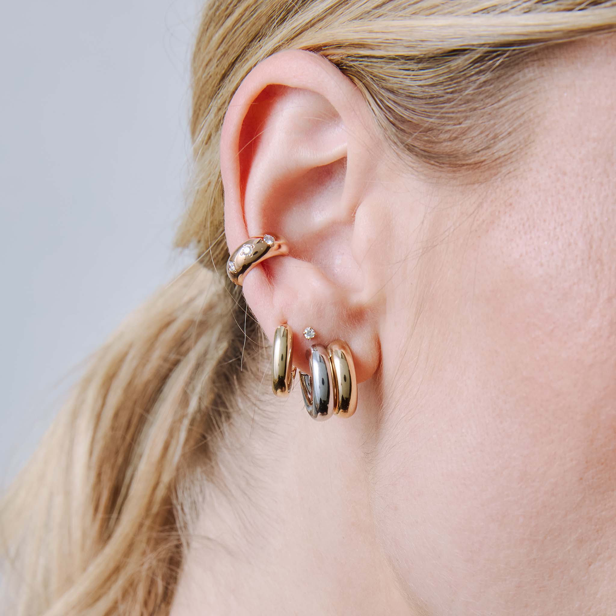 close up of a woman's ear wearing a Zoë Chicco 14k Gold Two Tone Gold Double Tube Huggie Hoop Earring layered with a 2mm Diamond Stud and a 14k Star Set Diamond Wide Chubby Ear Cuff