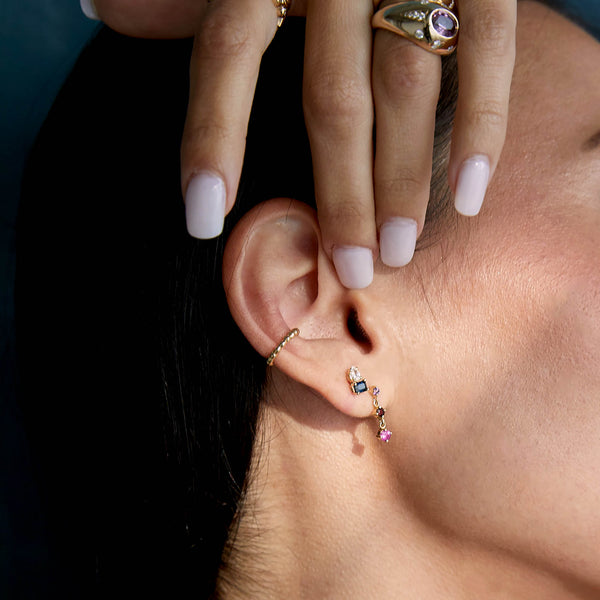 close up of a woman's ear wearing a Zoë Chicco 14k Gold Pear Diamond & Emerald Cut Blue Sapphire Stud Earring in her second piercing layered with a beaded ear cuff and an ombre pink sapphire gemstone drop earring