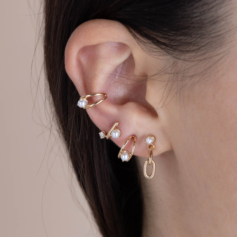 A woman's ear wearing a Zoë Chicco 14k Gold Mixed Prong Pearl & Diamond J Hook Hoop Earring in her third piercing layered with a 14k Pearl & Large Square Oval 2 Link Drop Earring, 14k Nested Pearl Double Wire Huggie Hoop, and a 14k Nested Pearl Double Wire Ear Cuff