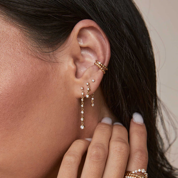 a close up of a woman's ear wearing a Zoë Chicco 14k Gold Mixed Prong Diamond Double Drop Earring in her second piercing and a 7 Linked Graduated Prong Diamond Drop Earring in her first piercing layered with two beaded ear cuffs