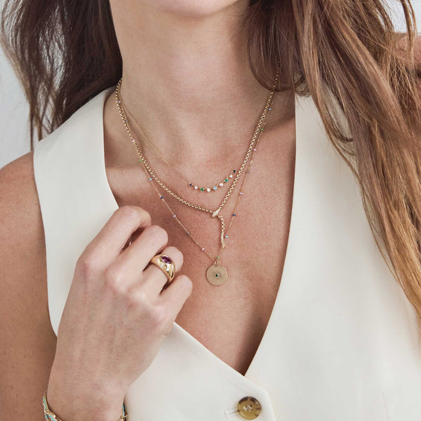 A woman in a beige vest wearing a Zoë Chicco 14k Gold Emerald Sunbeam Medallion Multicolor Satellite Chain Necklace layered with a Pavé Diamond Serpent Box Chain Necklace and a Linked Blue Ombre Gemstone Necklace