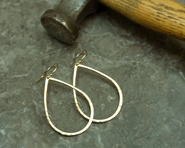 Zoe Chicco 14k gold teardrop hammered earrings on a textured surface with a hammer in the background