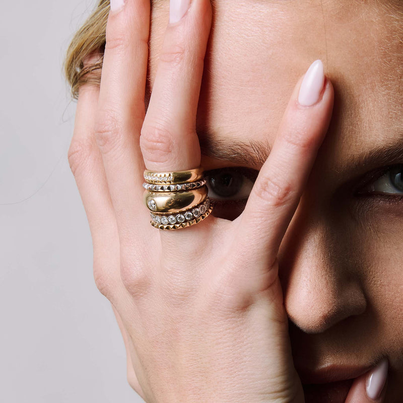 woman with her hand raised to her face wearing two Zoë Chicco 14k Gold Spike Eternity Rings in yellow and white gold stacked with a 14k 10 French Set Diamond Half Round Ring, Round Diamond Small Aura ring, and a 2.4mm Diamond Bezel Eternity Ring