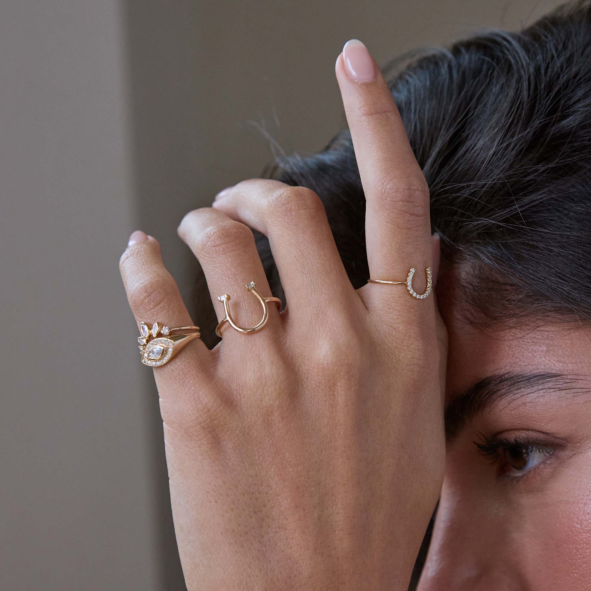 Close-up of a woman's hand wearing multiple Zoe Chicco 14k gold and diamond horseshoe shaped rings on a blurred grey backround