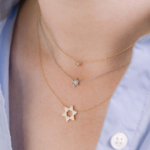 a close up shot of a woman in a blue shirt wearing a Zoë Chicco 14k Gold Itty Bitty Star of David Necklace layered with a 14k White Gold Midi Bitty Pavé Diamond Star of David Necklace and a Small Diamond Bezel Star of David necklace