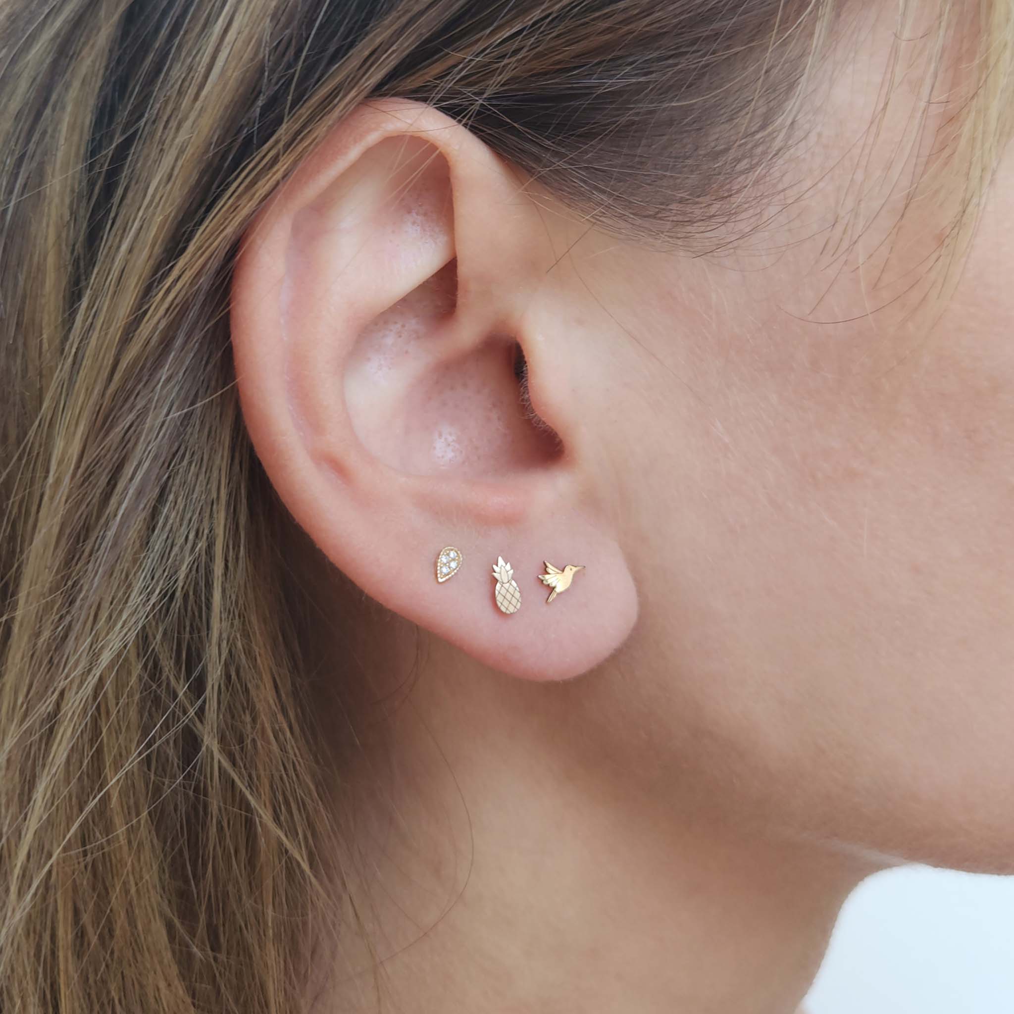 Close up of a woman's ear wearing a ZoÃ« Chicco 14k Gold Itty Bitty Pineapple Stud Earring in her second piercing and a 14k Itty Bitty Hummingbird Stud in her first piercing